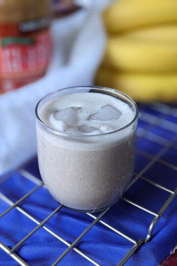 Peanut butter smoothie in glass with jar, cloth, and bananas in background